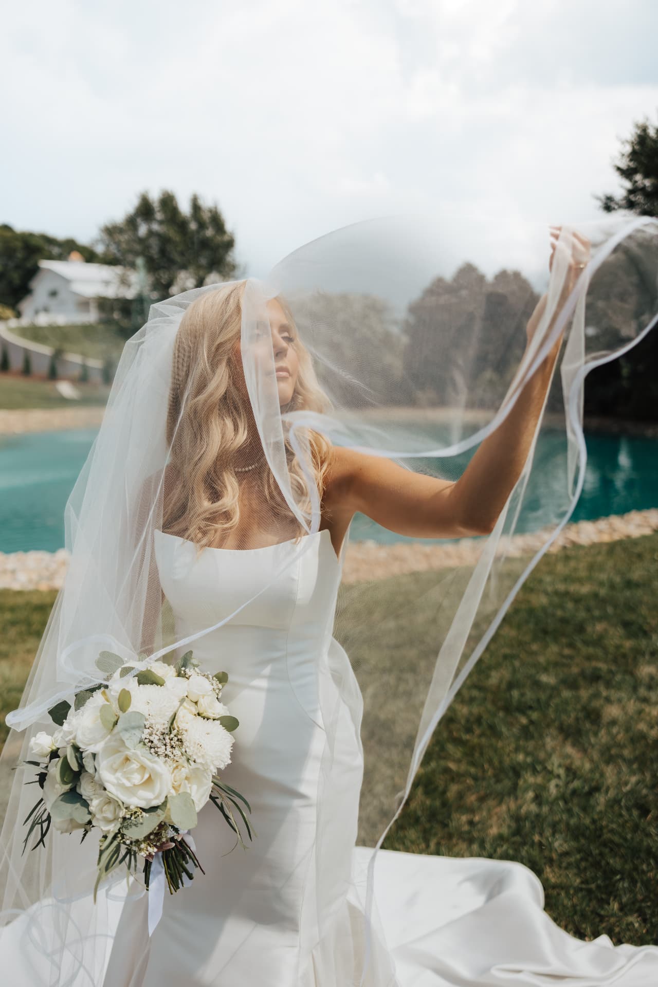 Bride lifting her veil into the wind beside a fountain pond