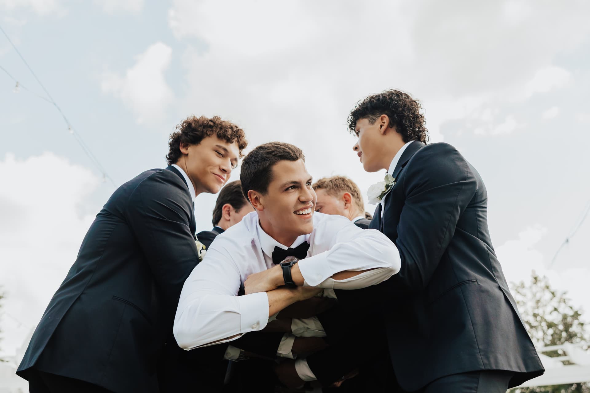 Groom lifted by groomsmen in tuxedos against a bright sky