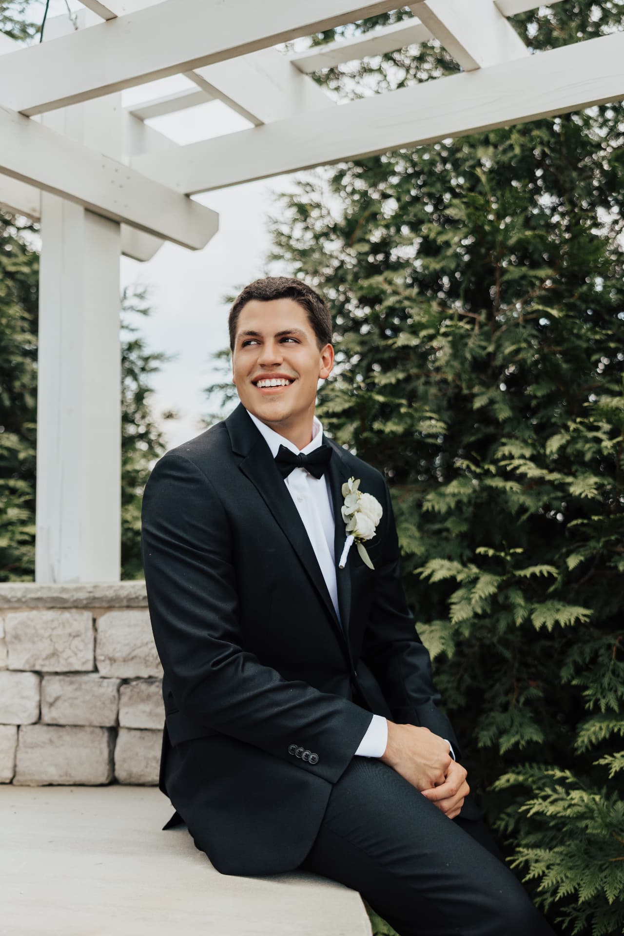 Groom seated beneath a white pergola smiling off camera