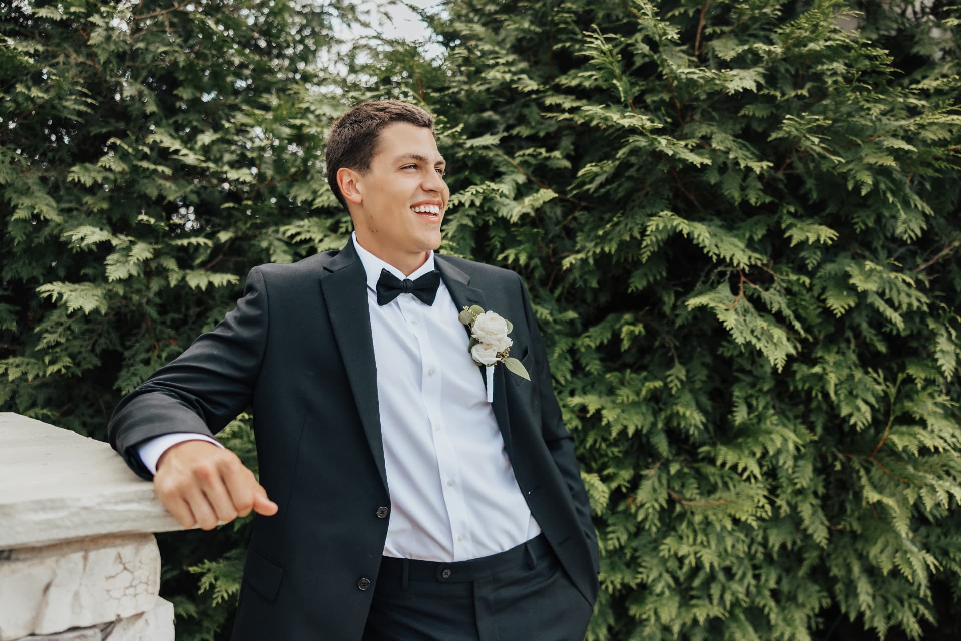 Groom in a black tuxedo leaning on stone against a green hedge