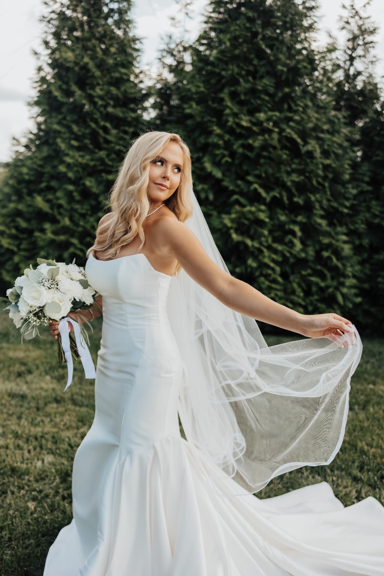 Bride turning with veil sweeping behind her against evergreens