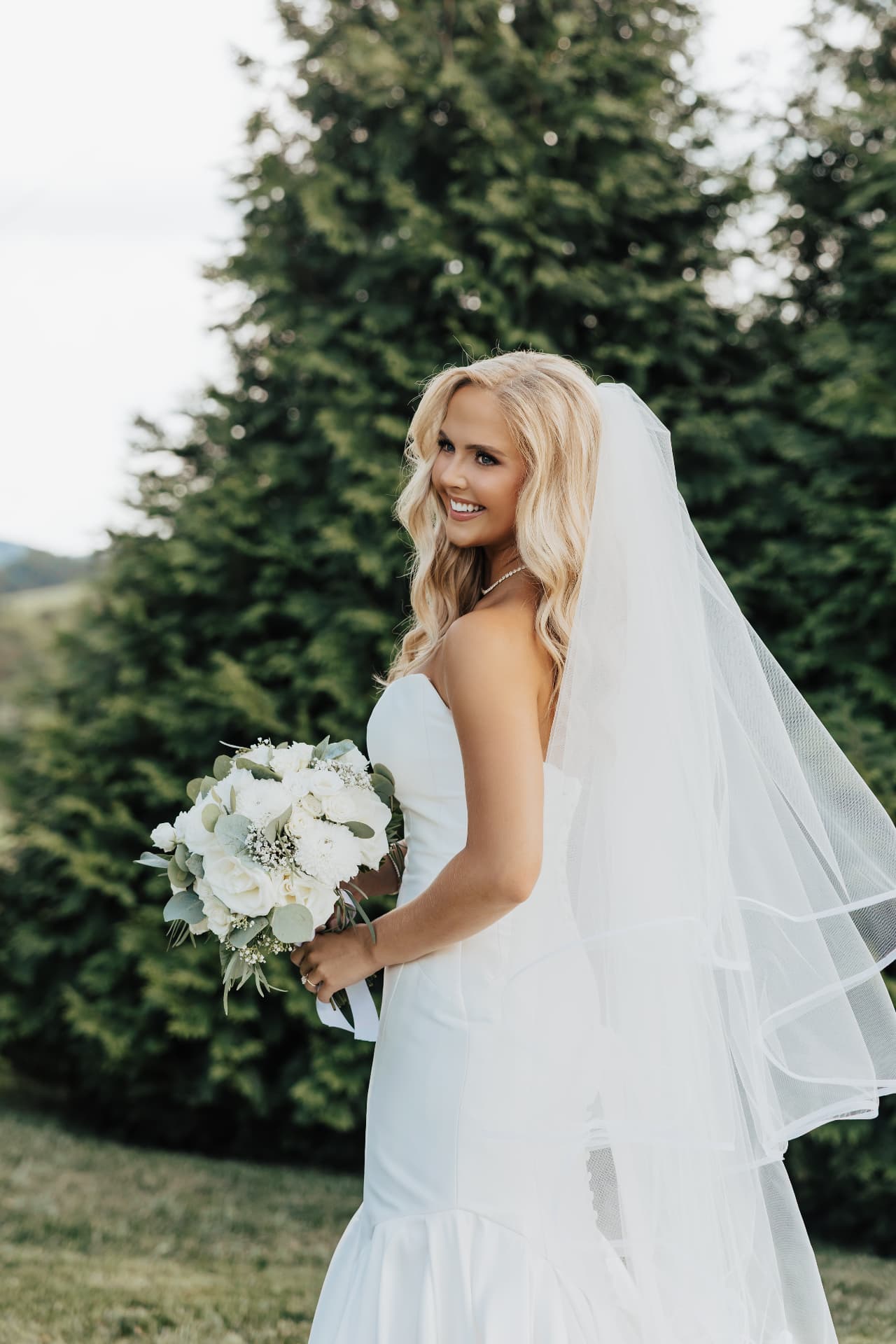Bride smiling in a strapless gown with veil against a pine backdrop